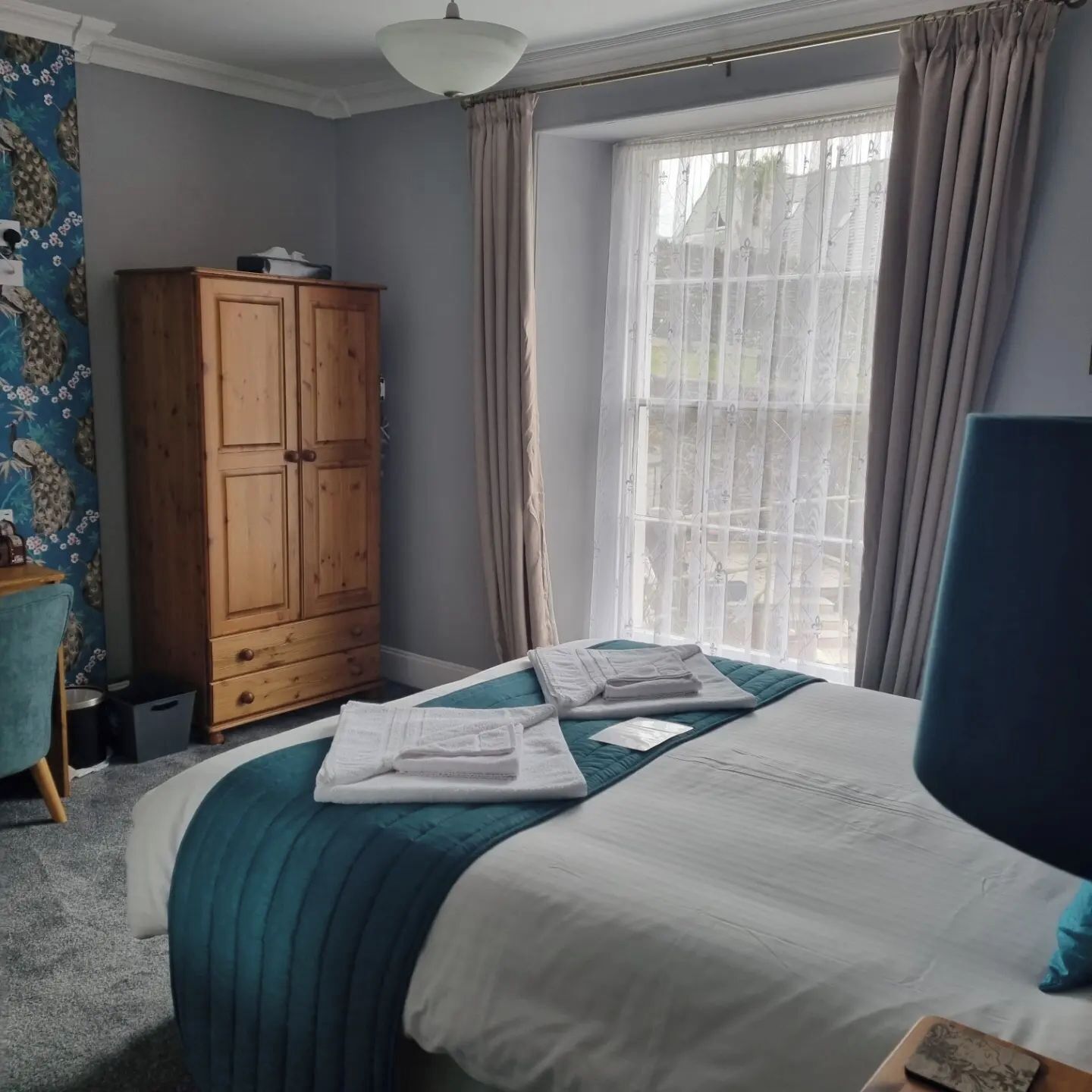 Una cama bien tendida con edredón verde agua y blanco, un armario de madera, una ventana con cortinas traslúcidas y una silla azul.