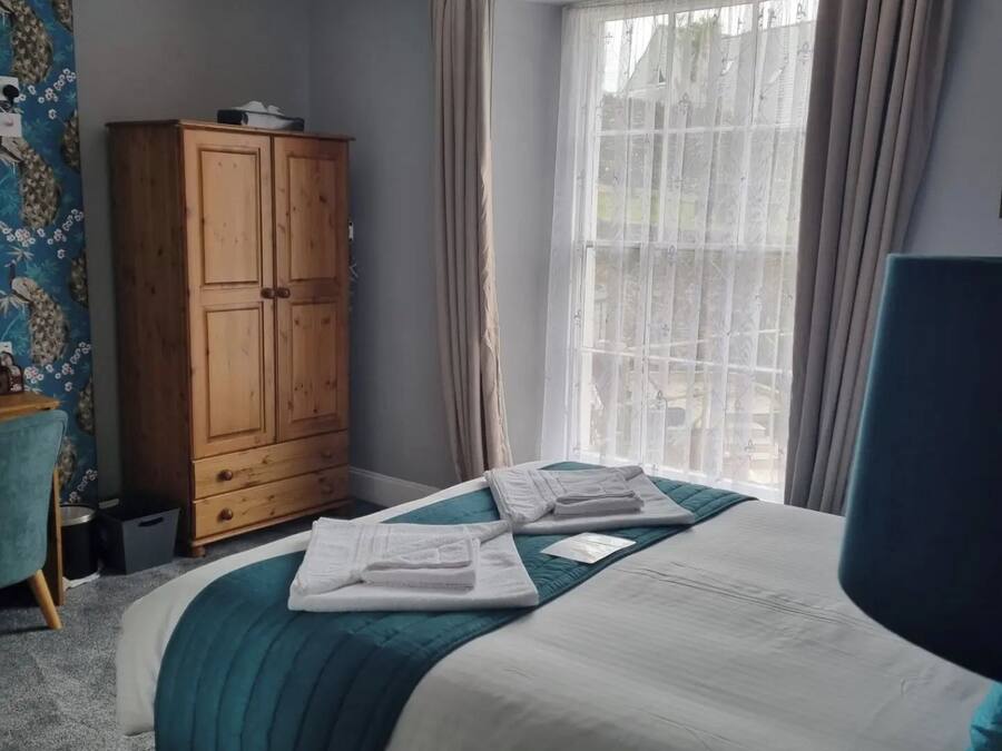 Una cama bien tendida con edredón verde agua y blanco, un armario de madera, una ventana con cortinas traslúcidas y una silla azul.