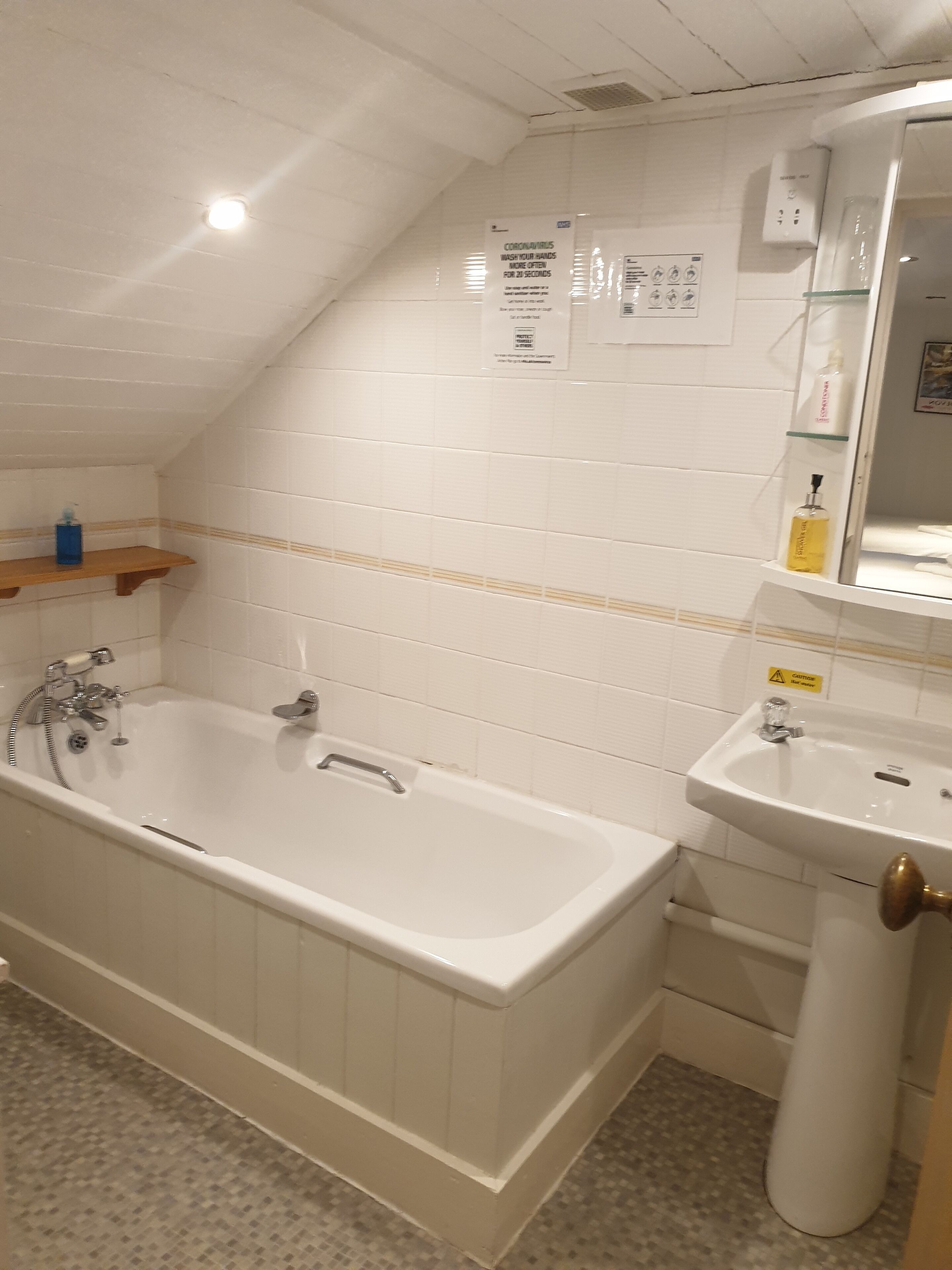 A bathroom with a bathtub, a sink, and a mirror. There are signs on the wall, a shelf with toiletries, and a tiled floor.