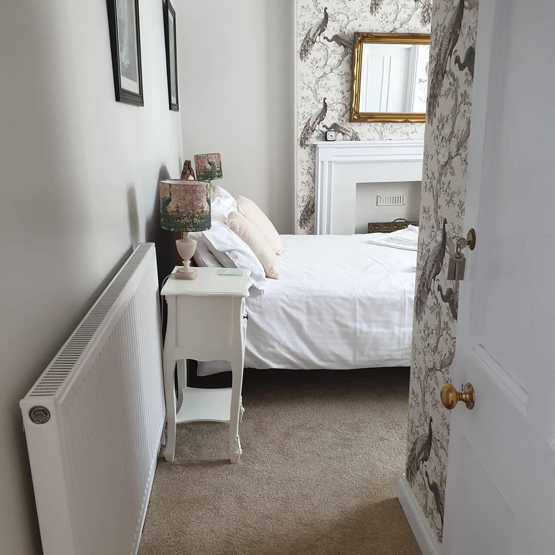 A bedroom with a white radiator, a small white nightstand, a bed with white bedding, a wall-mounted mirror, and a door with a floral wallpaper pattern.
