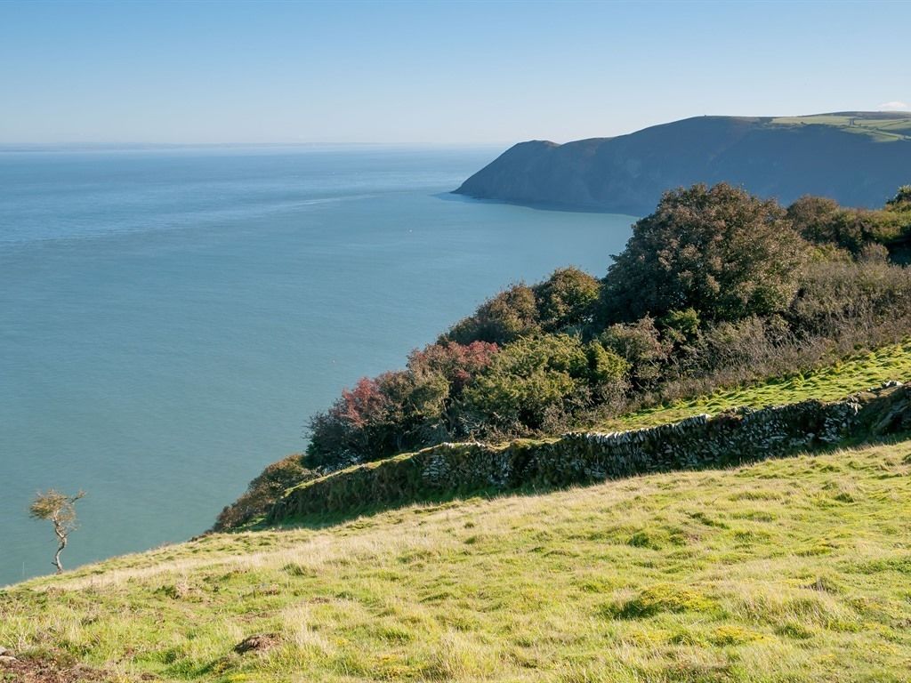Paisaje costero con un muro de piedra, terreno cubierto de hierba y vista al mar.