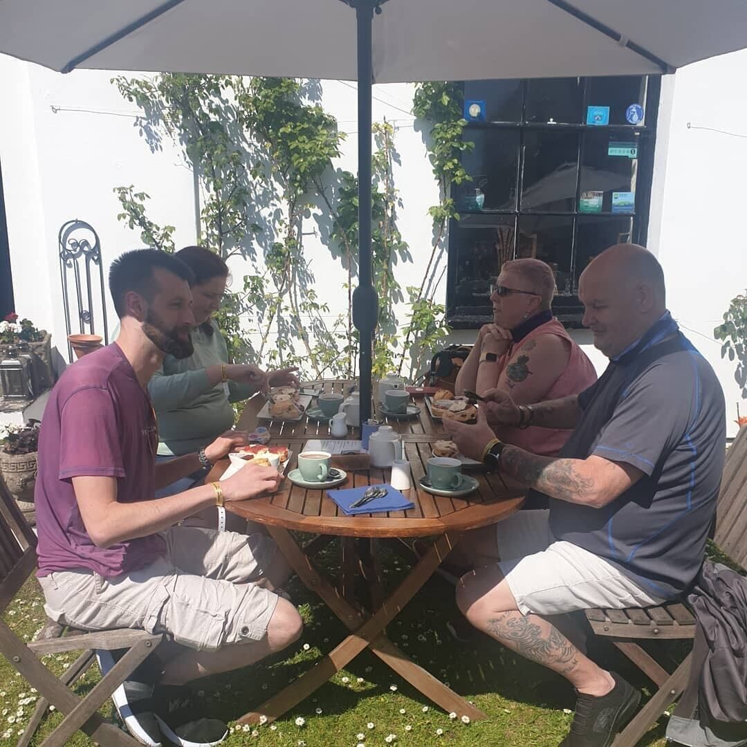 Um grupo de pessoas sentadas numa mesa redonda de madeira ao ar livre, com um guarda-sol branco acima.