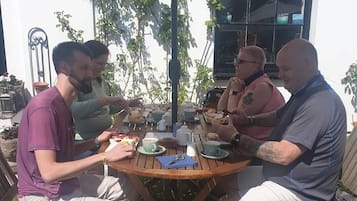 Um grupo de pessoas sentadas numa mesa redonda de madeira ao ar livre, com um guarda-sol branco acima.