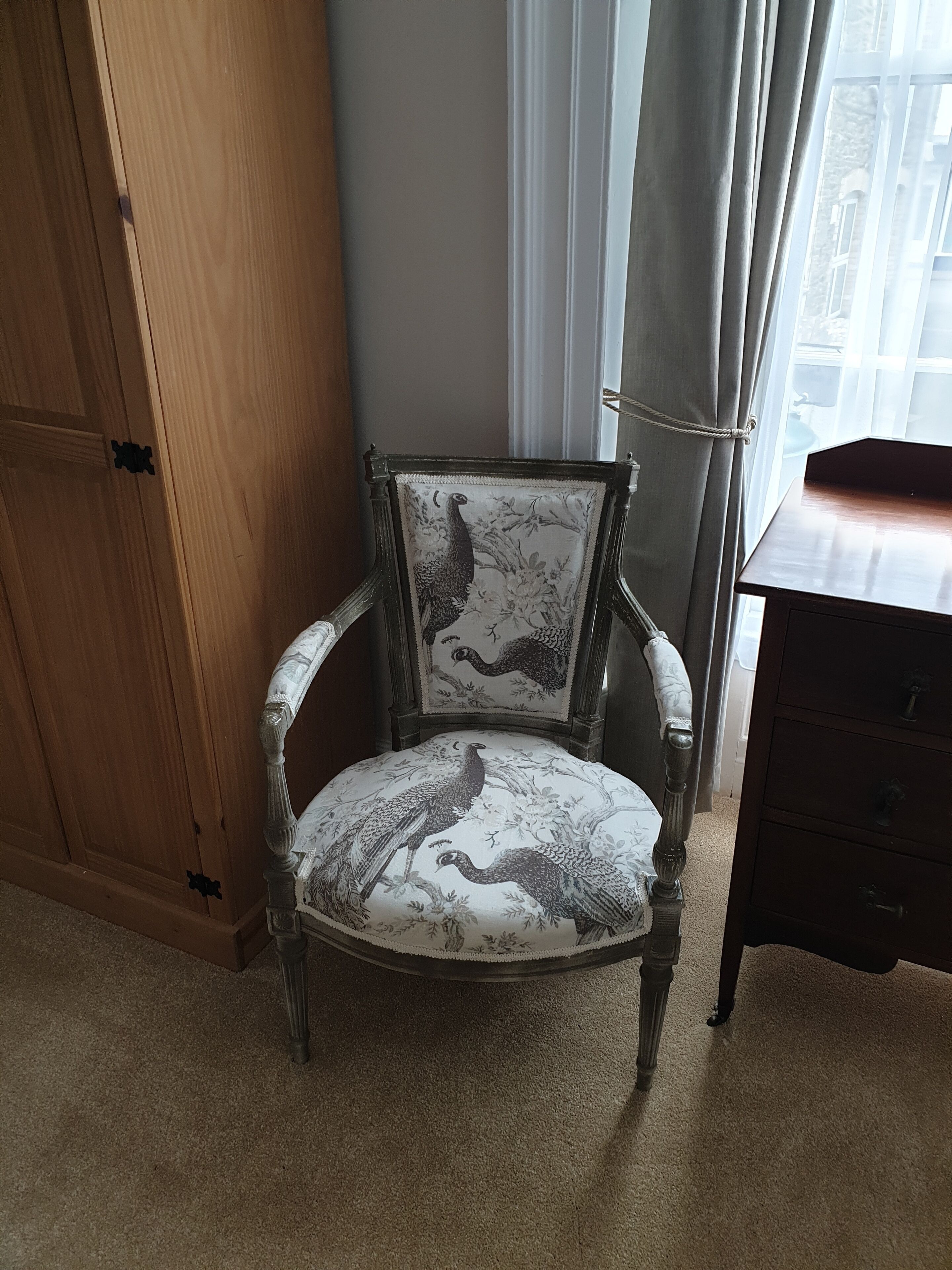 Una silla de estilo vintage con un diseño floral y de pavo real, ubicada en una habitación que incluye un armario de madera y un pequeño mueble de madera.