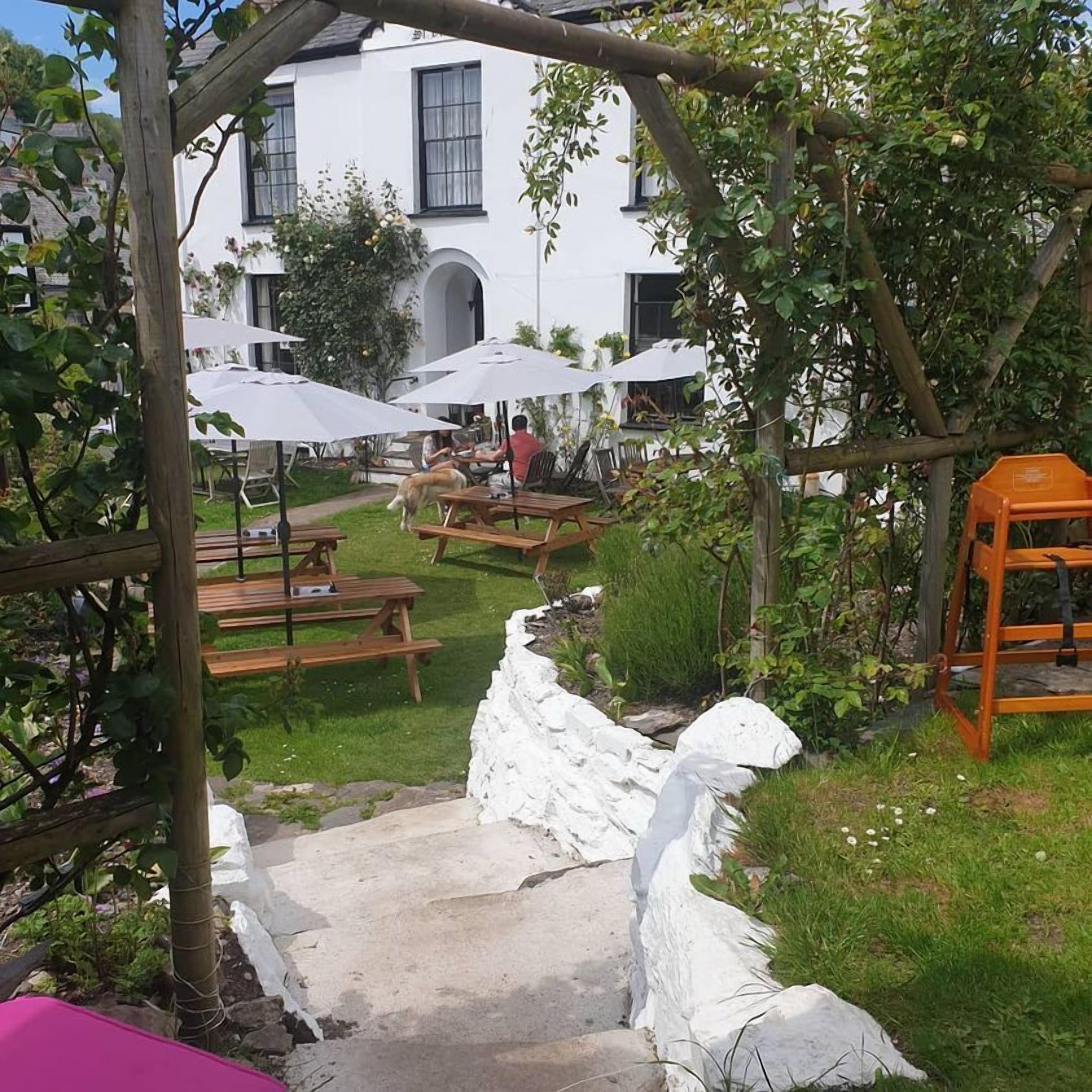 Un patio jardín con bancos de madera y sombrillas blancas, un muro de piedra y un edificio blanco al fondo.
