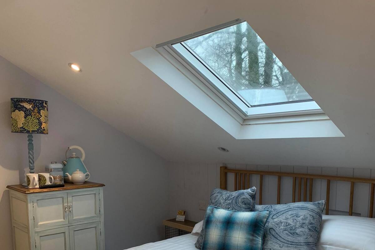 A bedroom with a skylight, a bed with white bedding, a wooden headboard, a nightstand with a lamp, and a dresser with decorative items.