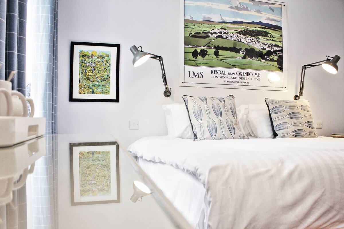 A neatly arranged bedroom with a bed, two bedside lamps, and framed maps on the wall.