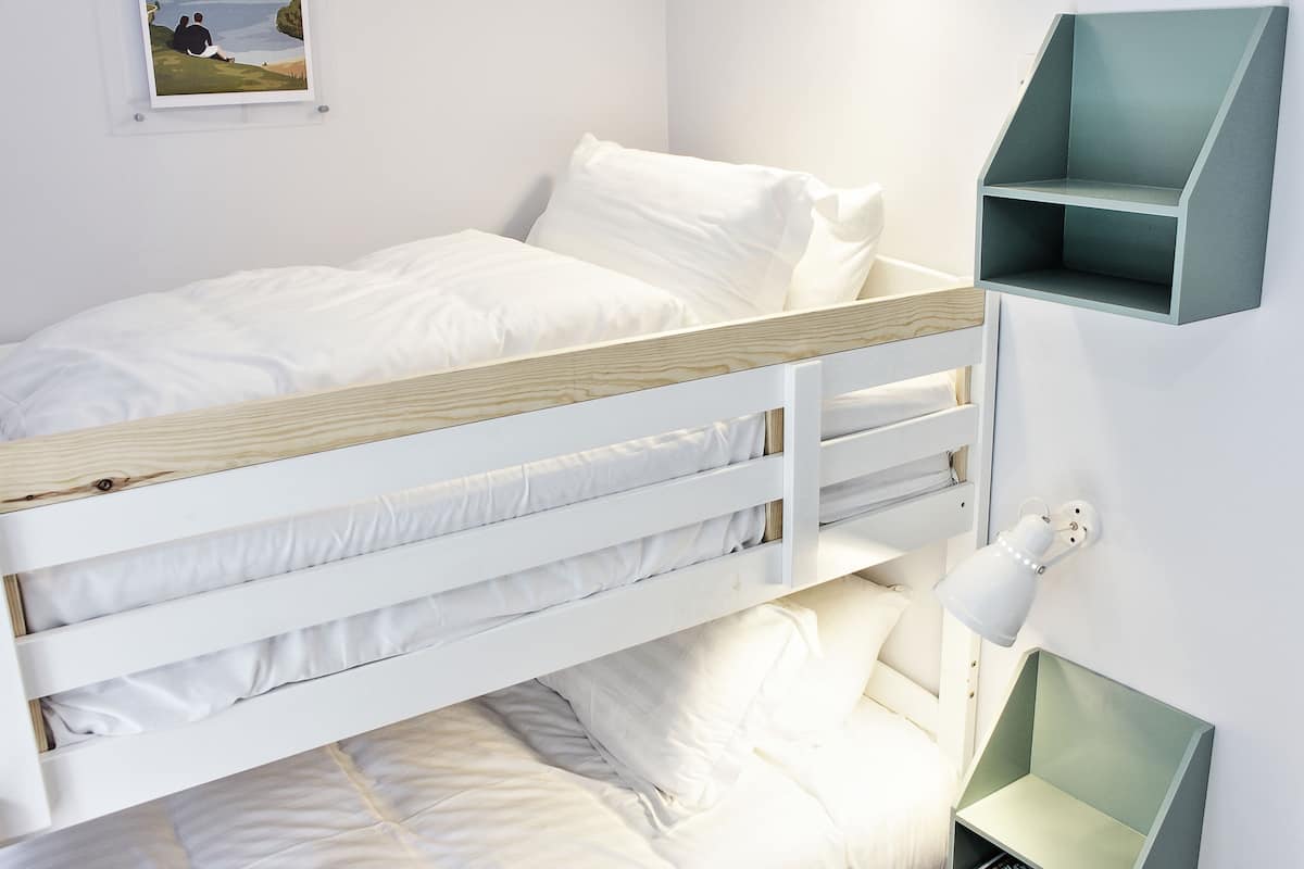 A bunk bed with white bedding, a small shelf unit, and a wall-mounted lamp.