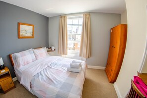 A neatly made bed with a checkered duvet, a bedside table with a lamp, and a window with curtains. - Olivers Guest House (Weymouth)