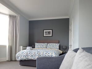 A bedroom with a bed, a grey sofa, a framed picture on the wall, and a window with curtains. - Olivers Guest House (Weymouth)