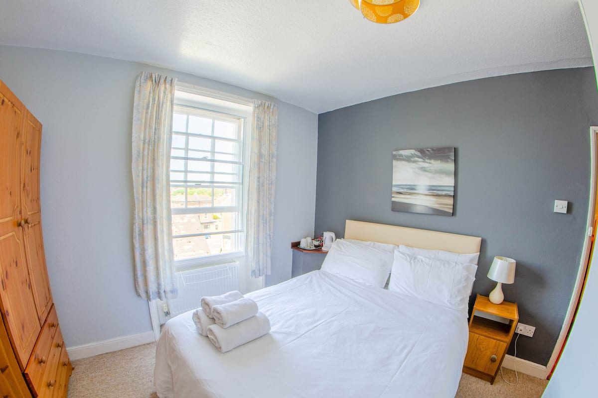 A neatly made bed with white linens and towels, a wooden nightstand with a lamp, a window with curtains, and a painting on the wall.