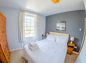 A neatly made bed with white linens and towels, a wooden nightstand with a lamp, a window with curtains, and a painting on the wall. - Olivers Guest House (Weymouth)