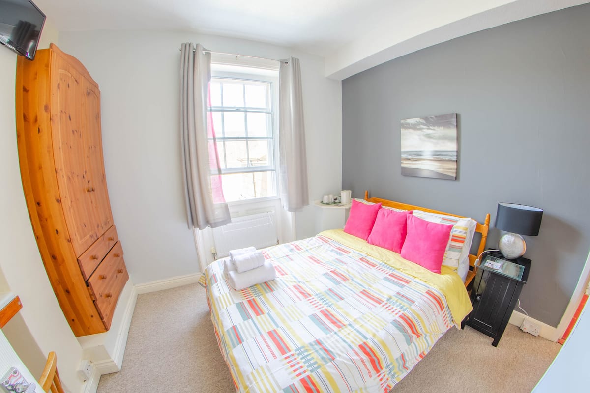 A bedroom with a bed, a window, a wooden wardrobe, a bedside table, and a wall-mounted lamp.