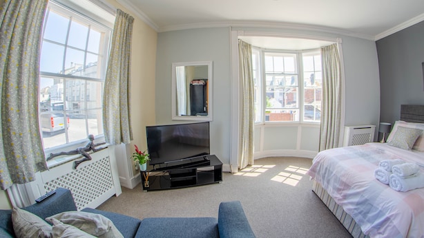 A hotel room with a large window, a flat-screen TV, a bed with pillows and towels, a sofa, and a radiator. - Olivers Guest House (Weymouth)