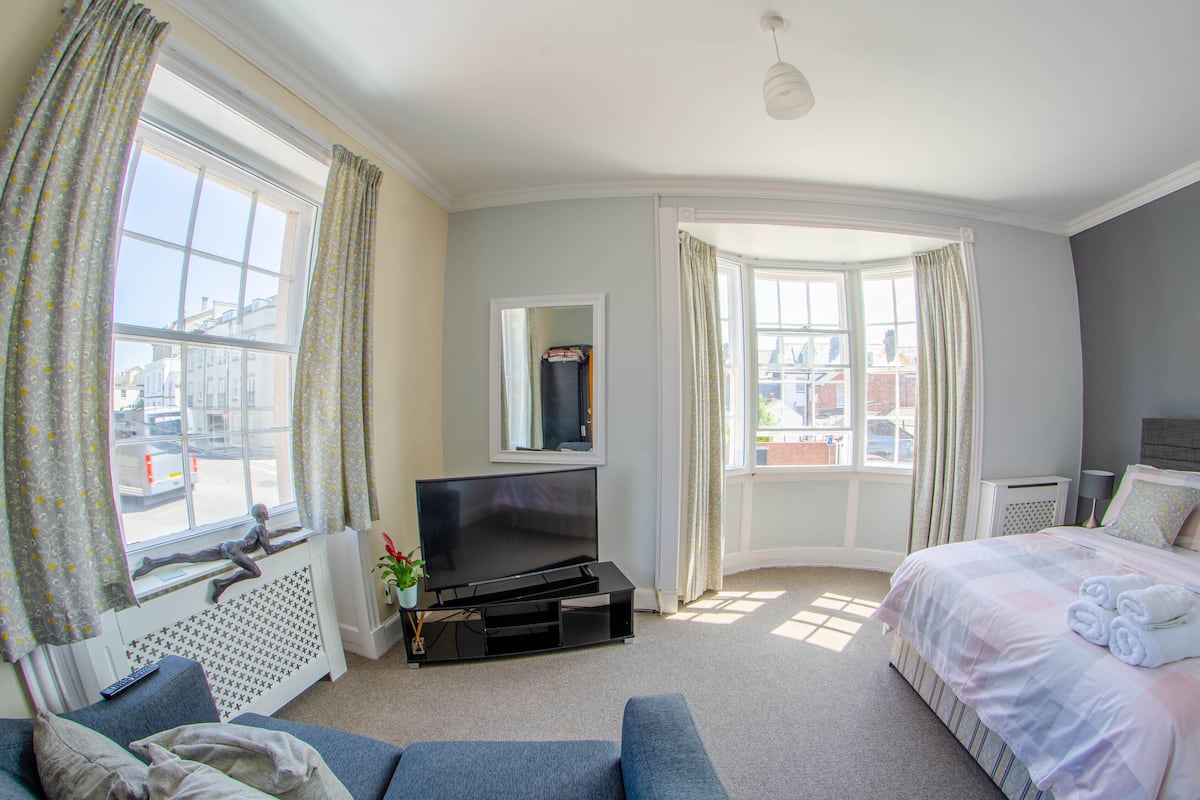 A hotel room with a large window, a flat-screen TV, a bed with pillows and towels, a sofa, and a radiator.