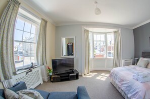 A hotel room with a large window, a flat-screen TV, a bed with pillows and towels, a sofa, and a radiator. - Olivers Guest House (Weymouth)