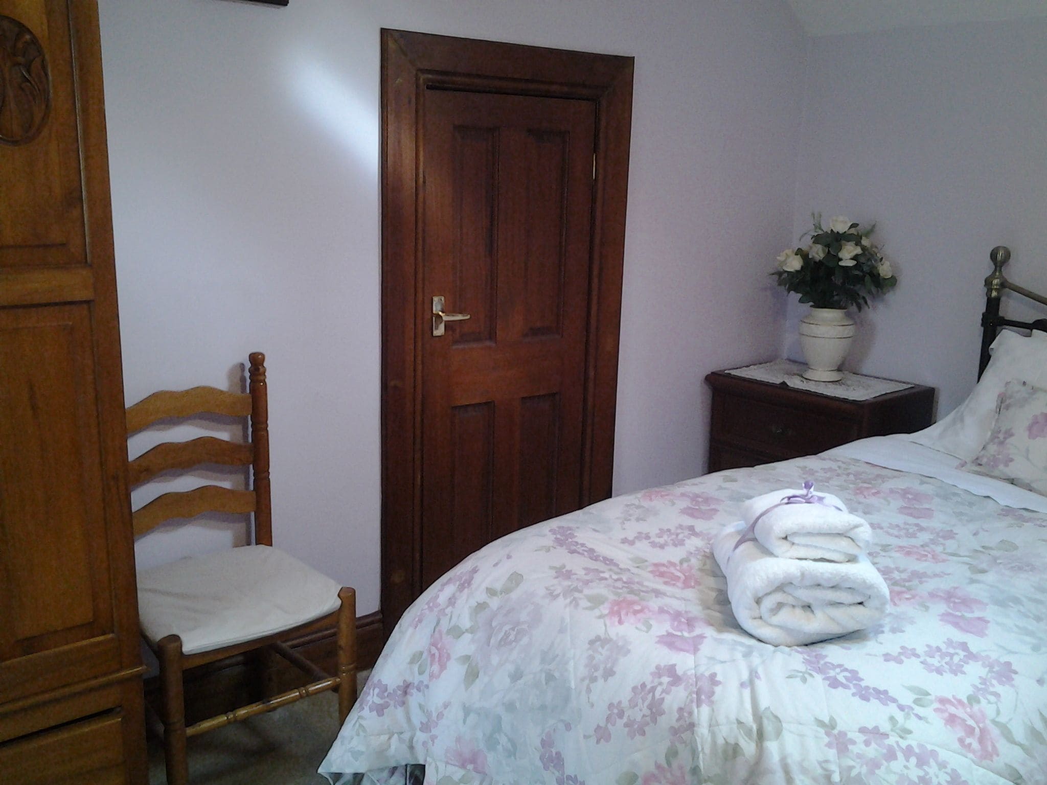A bedroom with a wooden chair, a bed with floral bedding, a nightstand with a potted plant, and a wooden wardrobe.