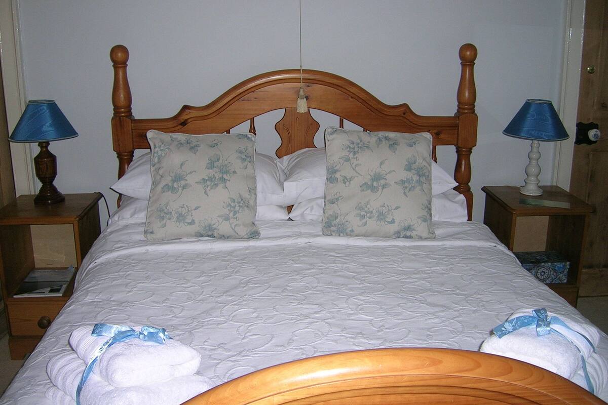 Una cama bien hecha con almohadones florales, cabecera de madera y dos mesitas de noche con lámparas.