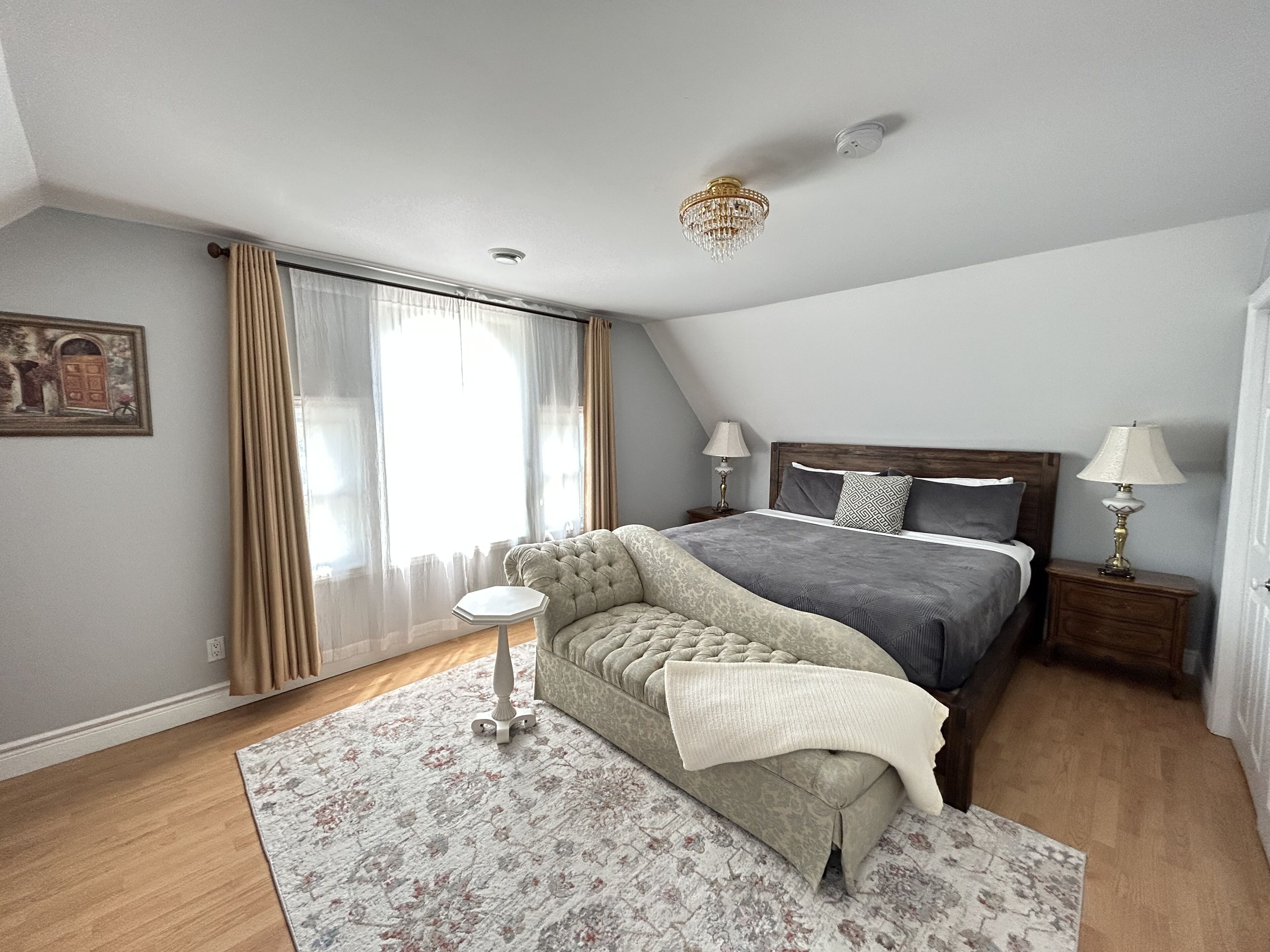 A bedroom with a bed, two bedside tables with lamps, a decorative wall piece, a window with curtains, and a patterned rug.