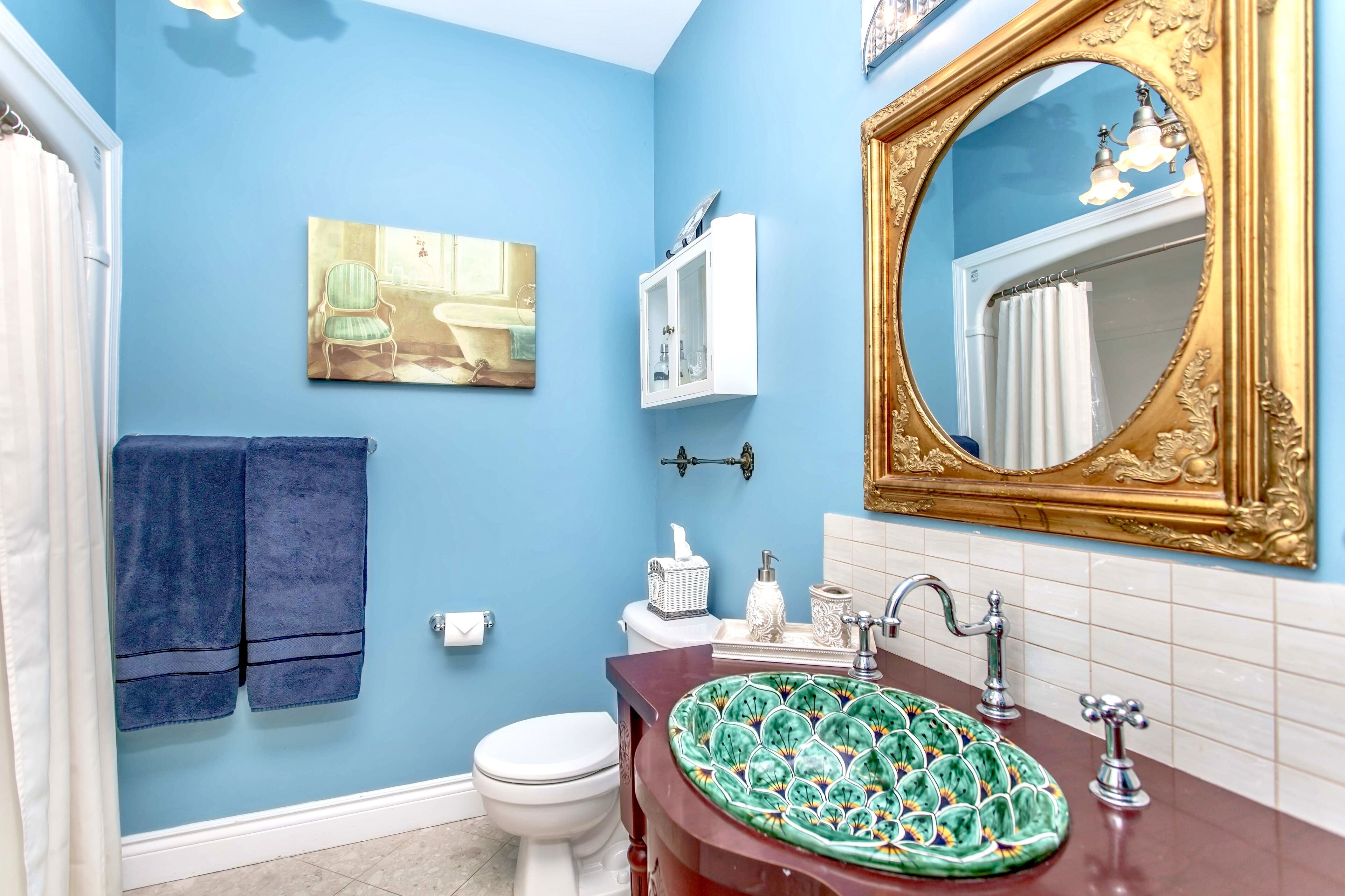 A bathroom with a blue wall, a white toilet, a green sink with a decorative bowl, a gold-framed mirror, and a framed picture above the sink.