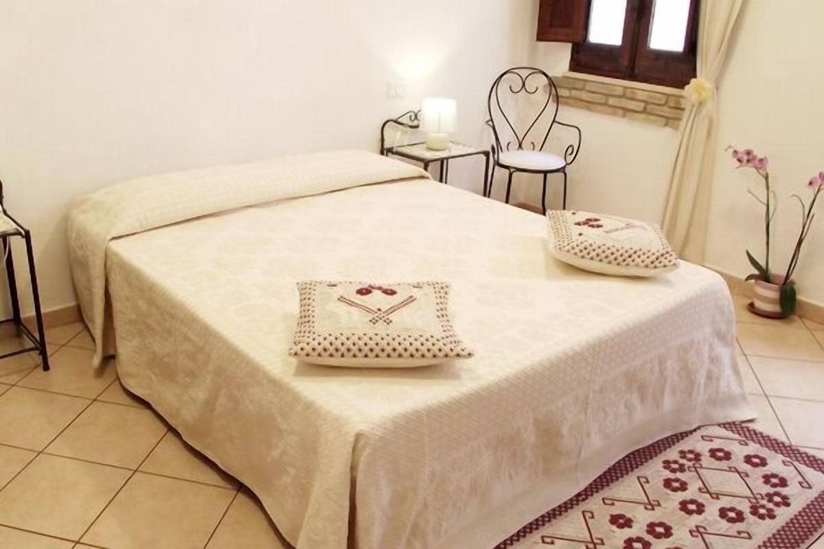 A neatly made bed with a beige bedspread and two decorative pillows, a small bedside table with a lamp, a chair with a heart-shaped backrest, and a potted plant.