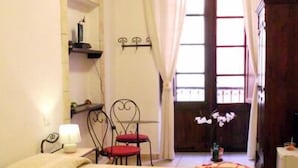 A room with two beds, a chair, a small table, a mirror, and a window with curtains. - Cagliari Castello Affittacamere (Cagliari)
