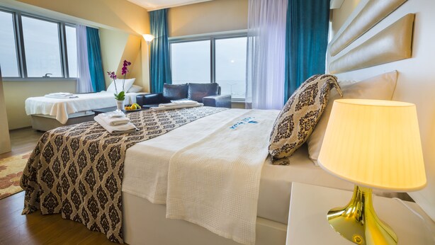 A hotel room with a large bed, a bedside table with a lamp, a chair, a small table with a vase of flowers, and a view of the city through large windows. - Arsen Hotel (Arsin)