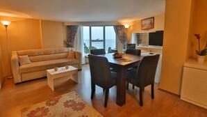 A compact living space with a sofa, dining table, chairs, a coffee table, a TV, and a view of the sea. - Arsen Hotel (Arsin)