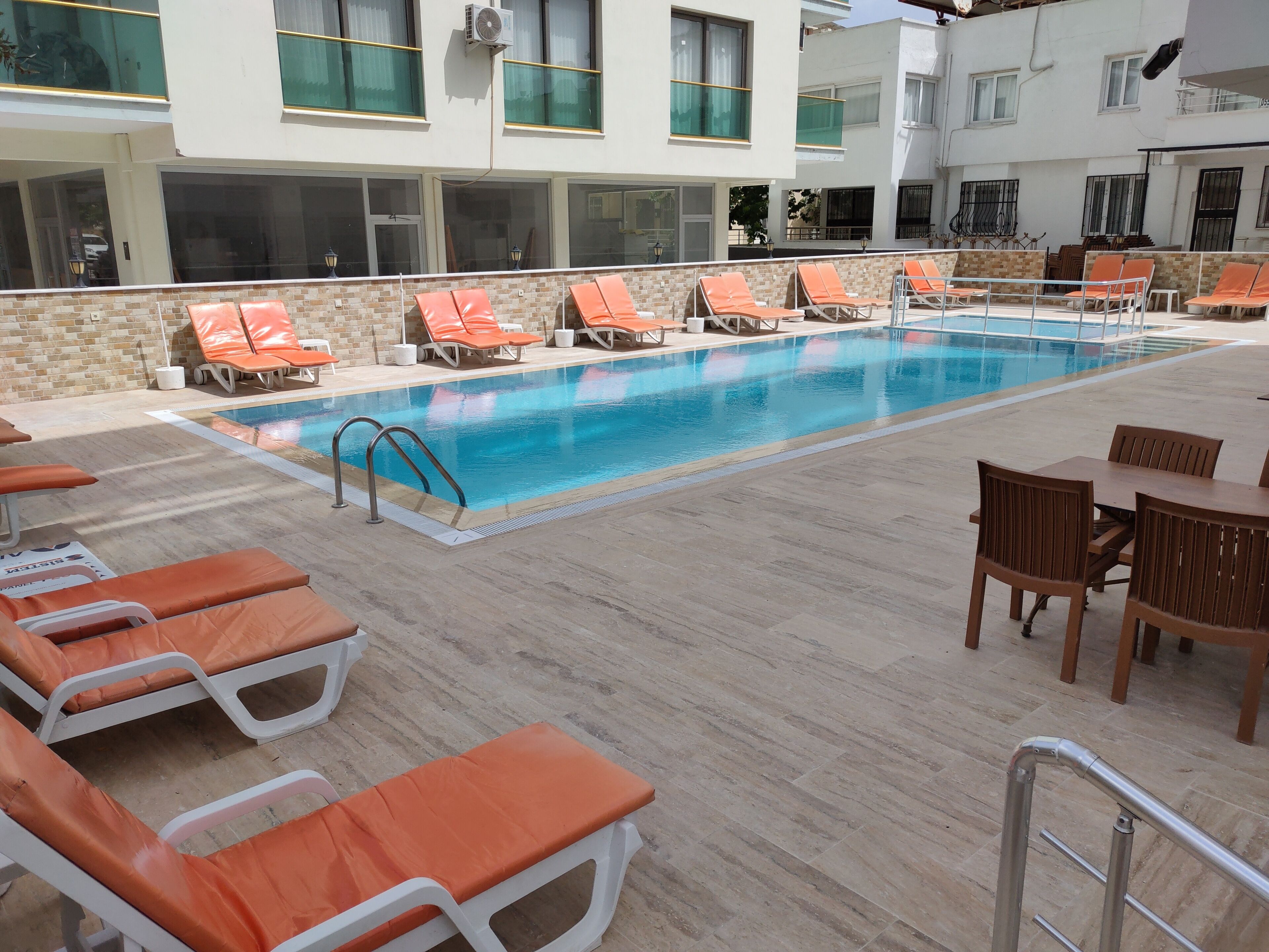 A pool area with lounge chairs and a table, surrounded by a tiled deck and a building with multiple windows.