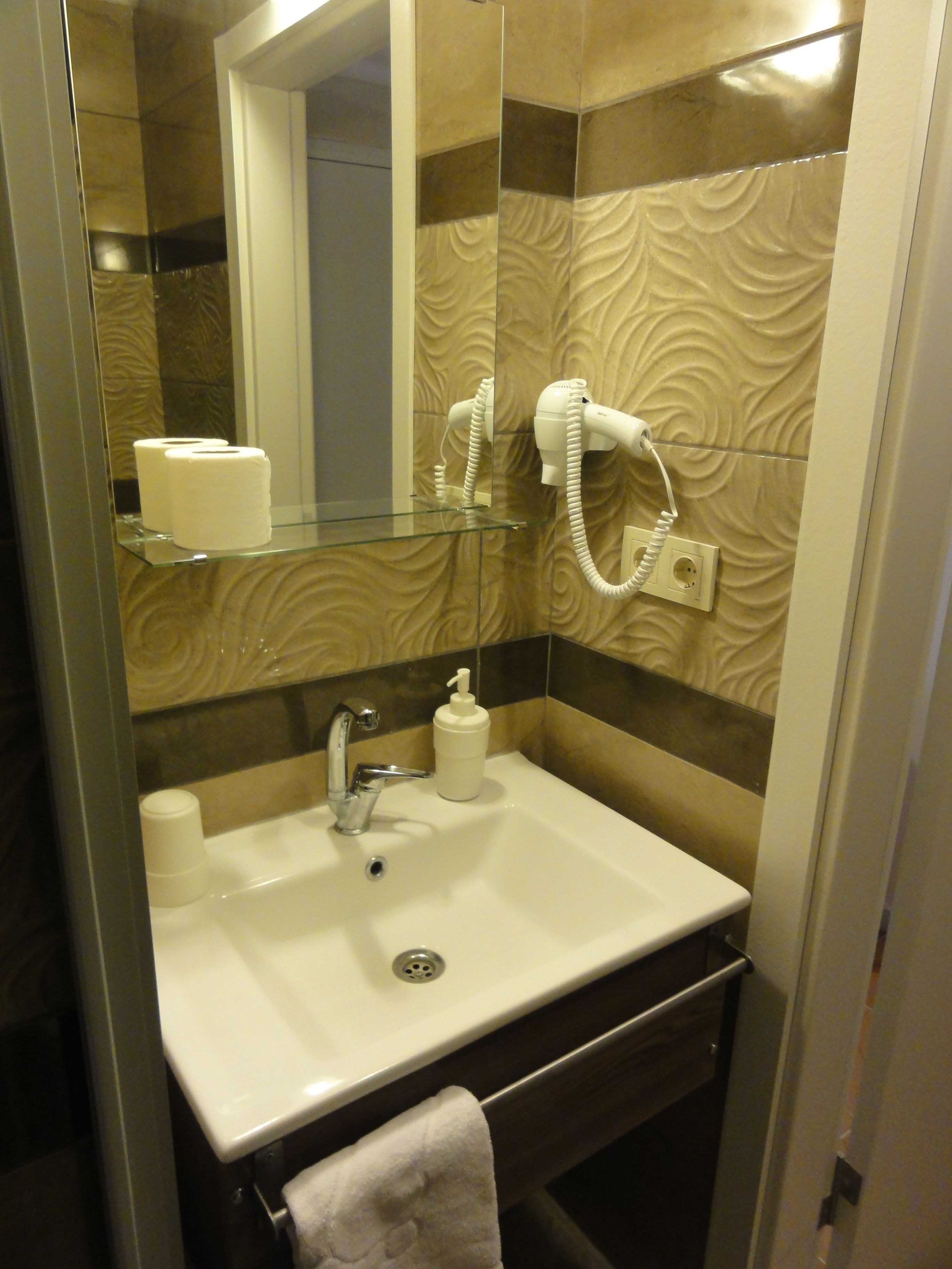 A bathroom with a glass shelf holding toilet paper, a white sink with a faucet, a hand soap dispenser, and a towel hanging on a rack.