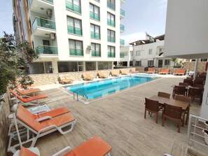 Uma área de piscina com espreguiçadeiras e mesas, rodeada por um edifício com vários andares e varandas.