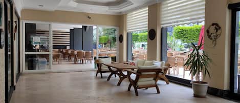 A spacious indoor area with wooden tables and chairs, large windows with blinds, and a view of the outside.