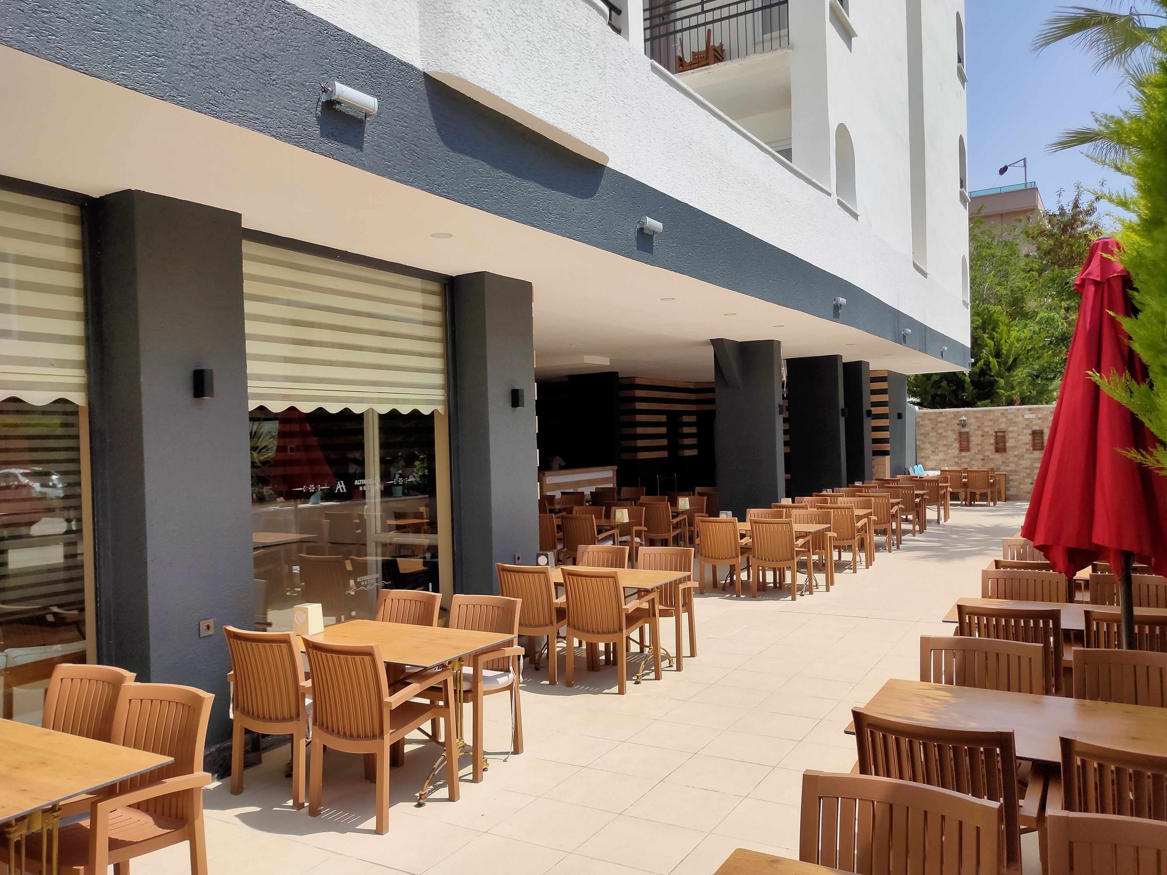 Un espace extérieur aménagé en terrasse, avec plusieurs tables et chaises en bois, un parasol rouge et une vue sur un immeuble doté d’un balcon.