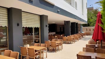 Un patio al aire libre con varias mesas y sillas de madera, un paraguas rojo y vista a un edificio con balcón.