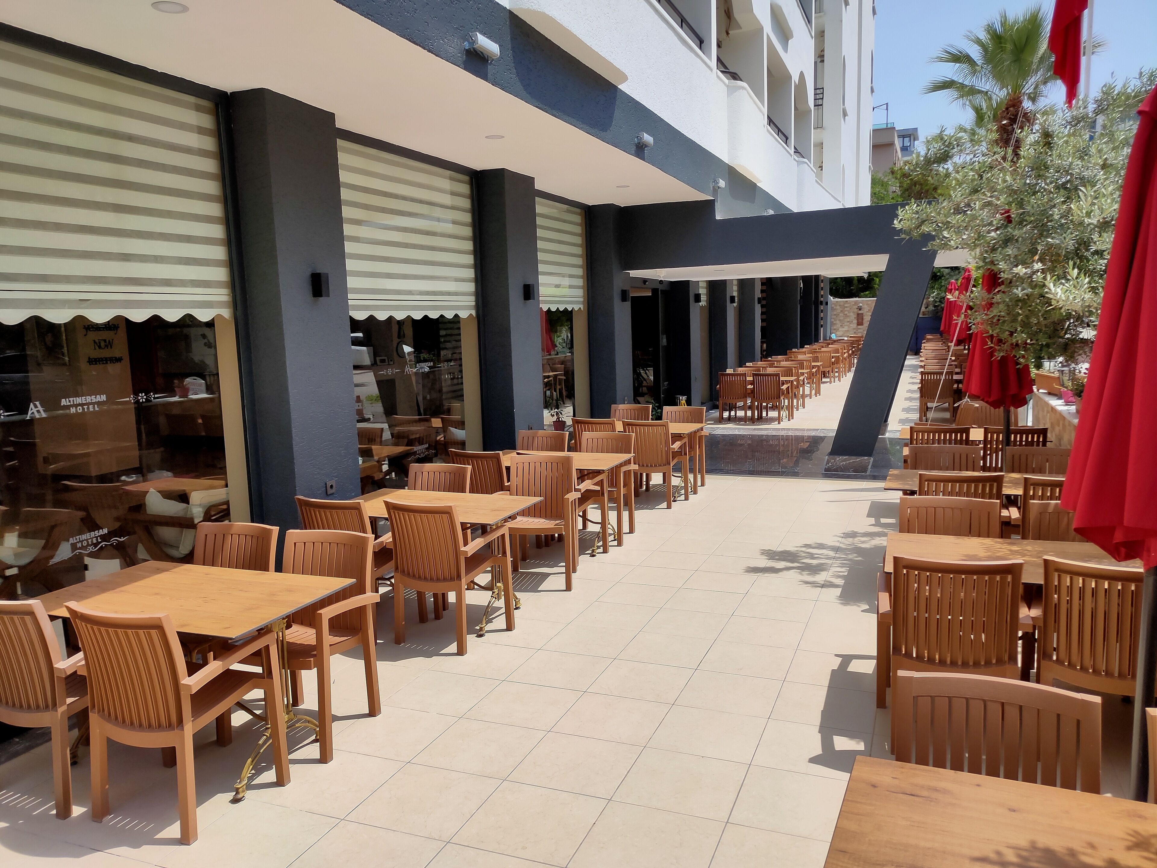 Un espace café en plein air avec des tables et des chaises en bois, un sol carrelé et des parasols rouges.