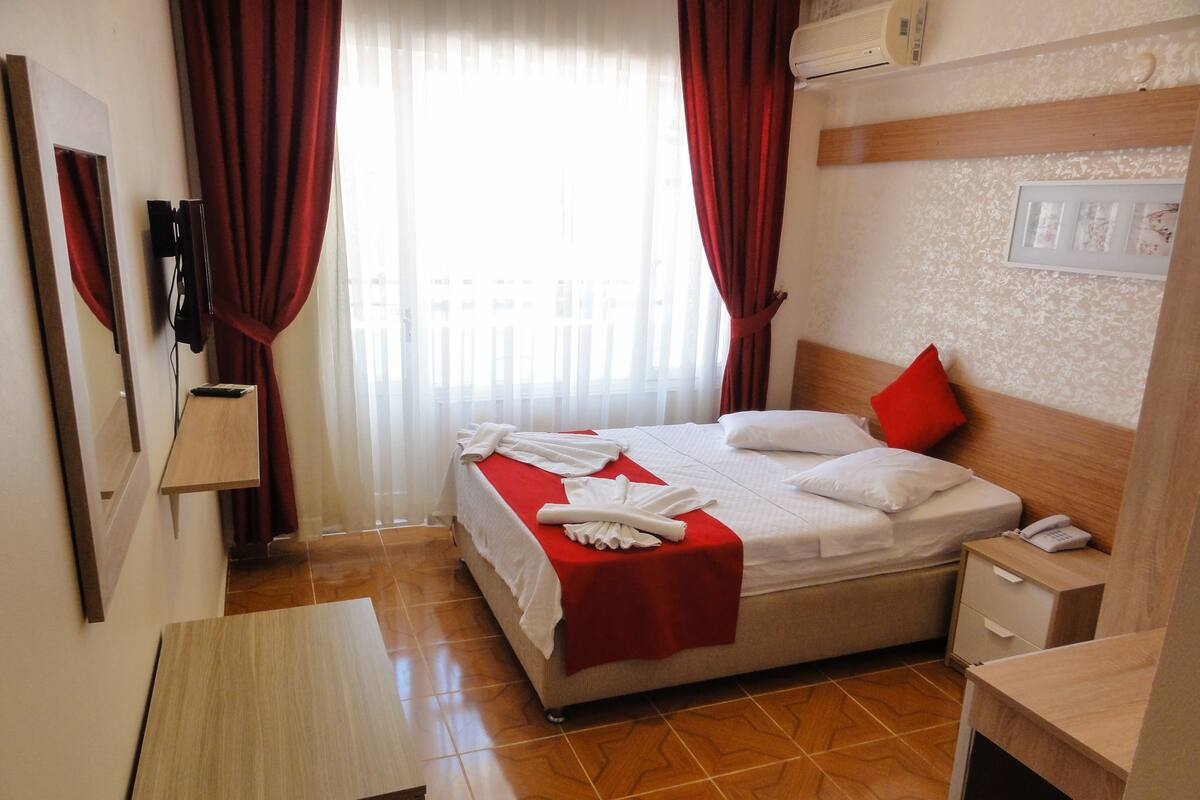 A hotel room with a bed, a bedside table, a nightstand, a wall-mounted phone, and a window with curtains.