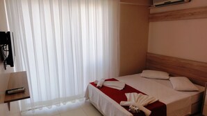 Ein Hotelzimmer mit Bett, Holz-Kopfteil, rotem Akzent auf dem Bett, weißem Vorhang, Wand-Klimaanlage und einem kleinen Holztisch mit Fernbedienung.