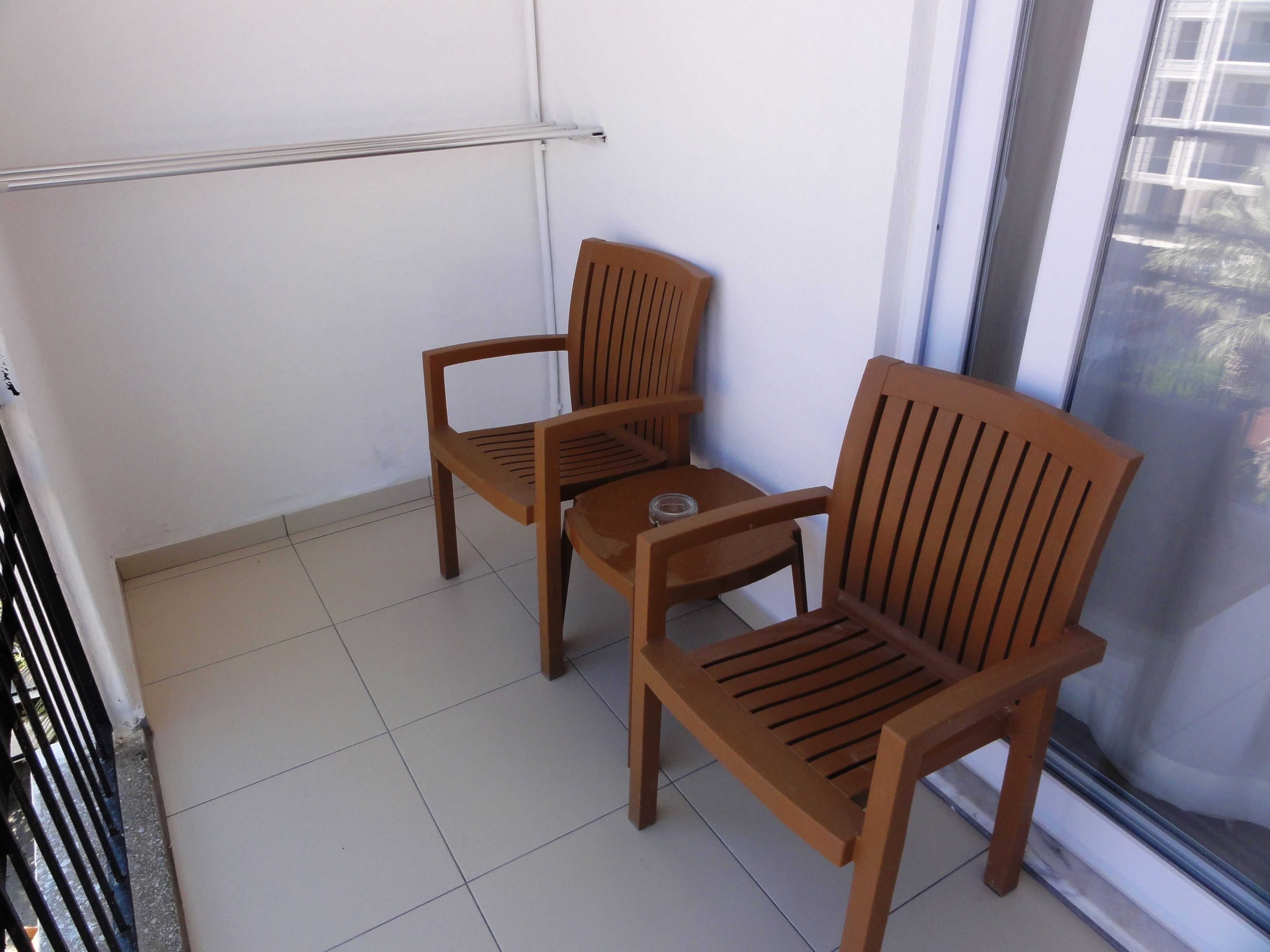 A balcony with two wooden chairs and a small table, a metal railing, and a view of buildings through a glass door.