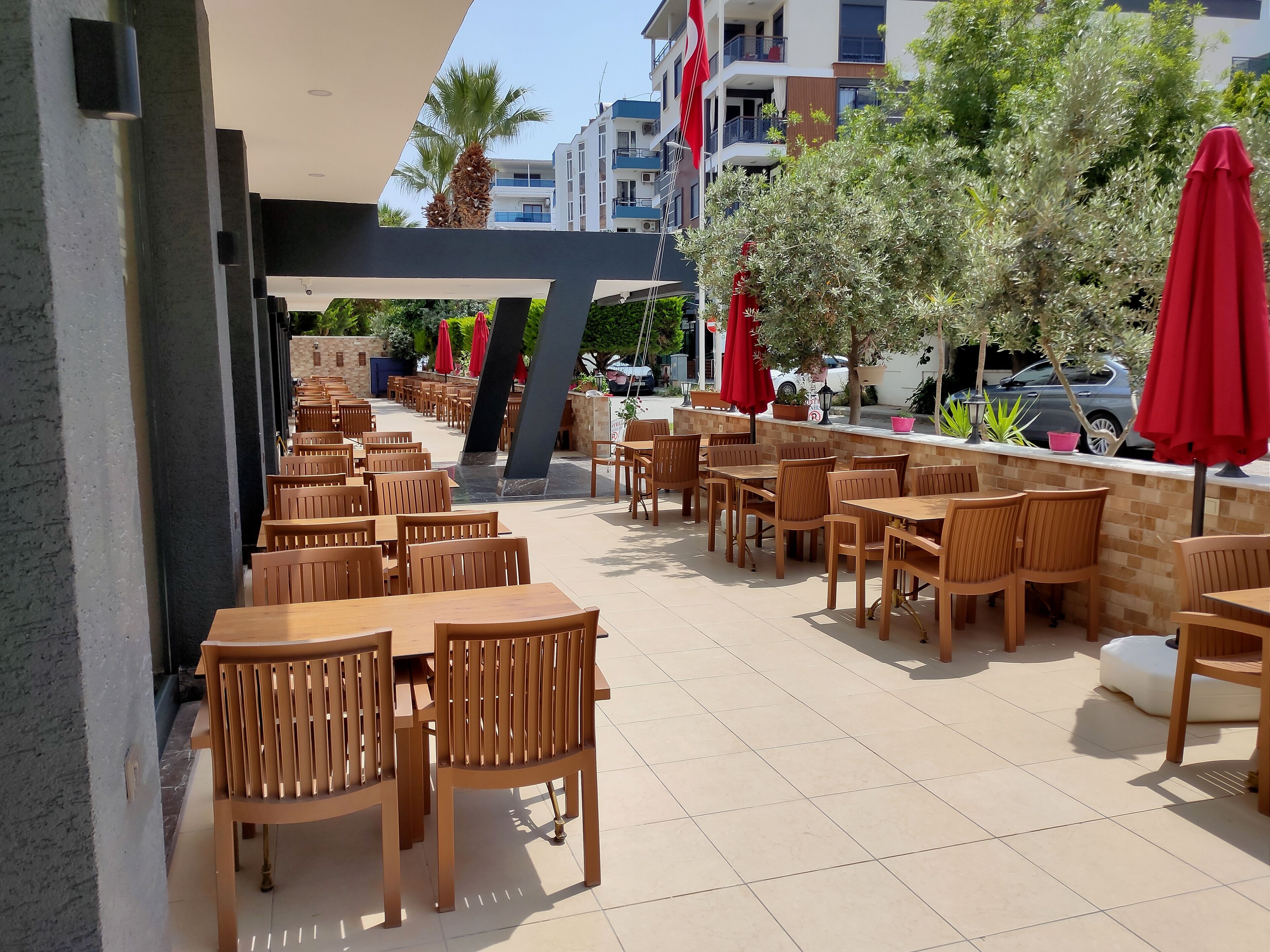 An outdoor cafe area with wooden tables and chairs, red umbrellas, and a view of buildings in the background.
