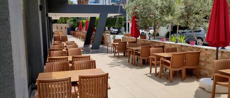 An outdoor cafe area with wooden tables and chairs, red umbrellas, and a view of buildings in the background.