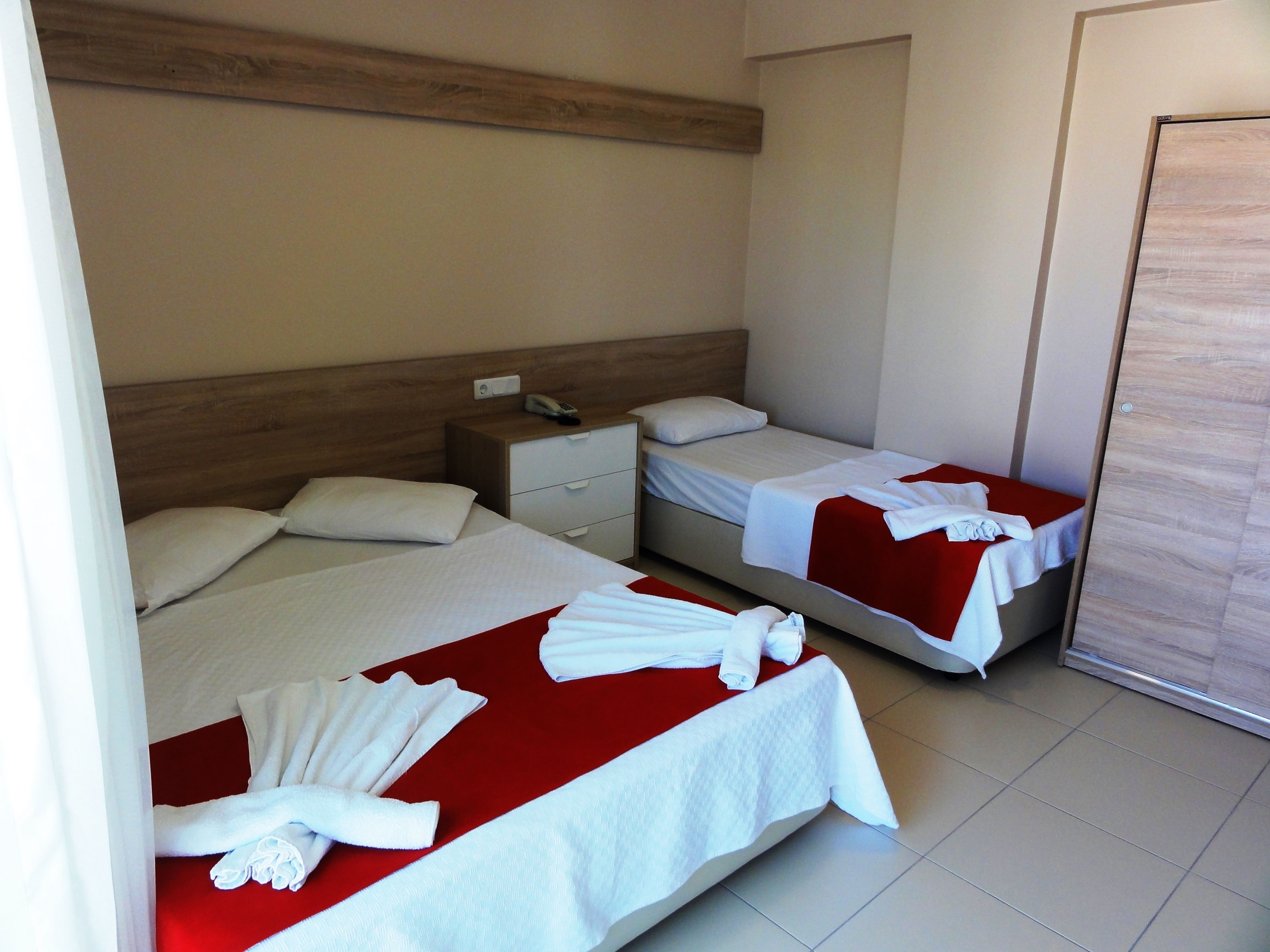 Een hotelkamer met twee bedden, elk met een rode spreideken en witte handdoeken. Er is een houten hoofdeinde, een nachtkastje met een telefoon en een deur aan de rechterkant.