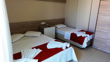 Een hotelkamer met twee bedden, elk met een rode spreideken en witte handdoeken. Er is een houten hoofdeinde, een nachtkastje met een telefoon en een deur aan de rechterkant.