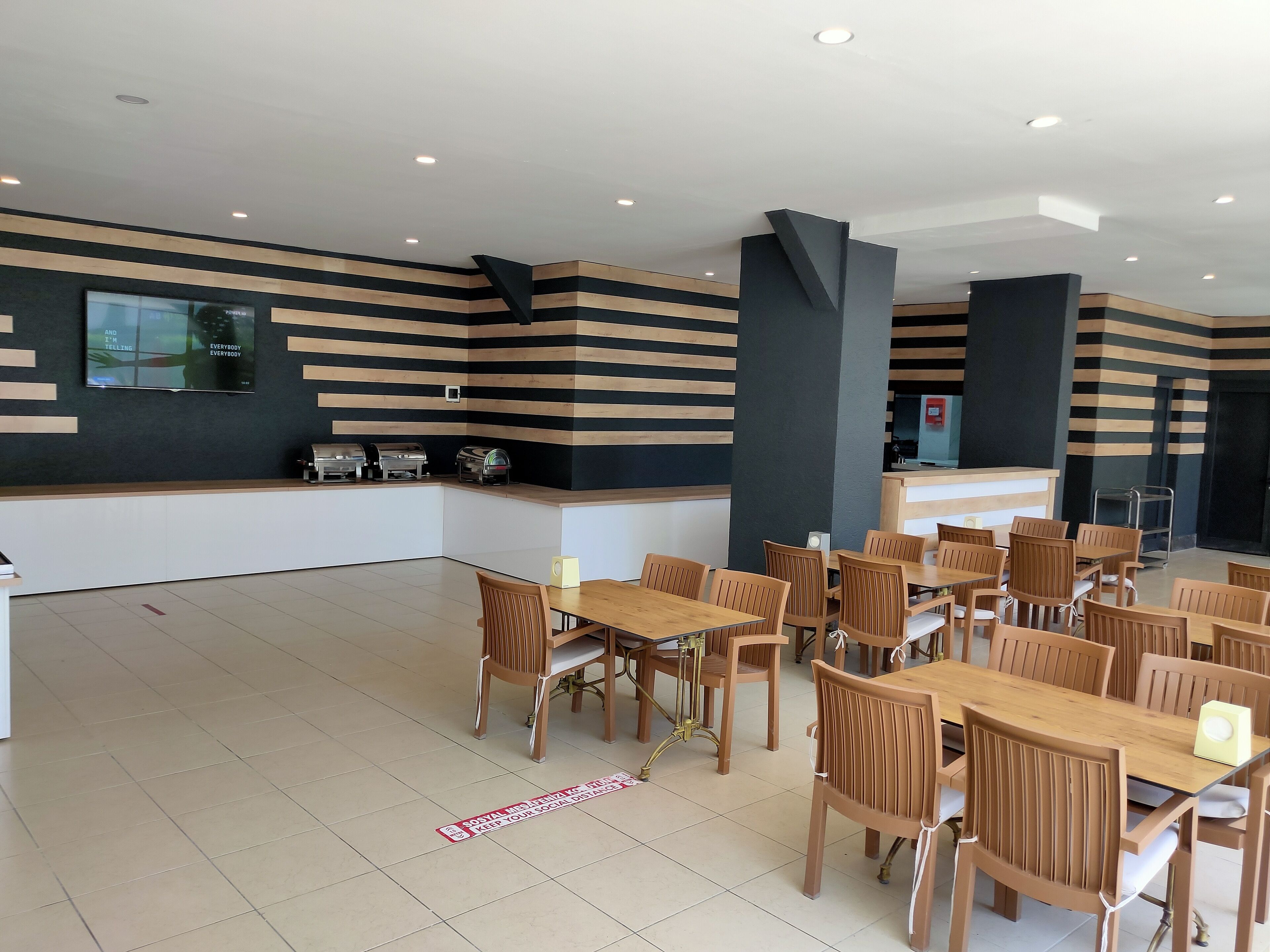 A modern dining area with wooden tables and chairs, a counter with food service equipment, and a large screen on the wall.