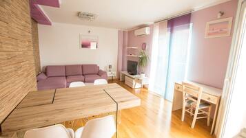 A modern living room with a wooden dining table, white chairs, a purple sofa, a TV, and a small desk with a chair.