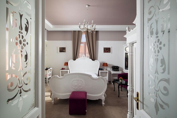 Une chambre à coucher avec un lit blanc à baldaquin, un lustre, un bureau avec une machine à écrire et un pouf violet.
