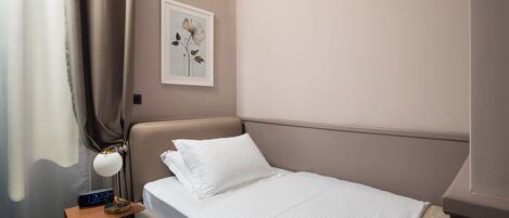 Una cama bien hecha con ropa blanca, una lámpara de noche y un reloj digital sobre una mesita de noche.