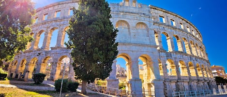 An ancient amphitheater with multiple arches and a central stage, surrounded by trees and a paved walkway.
