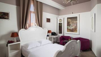 A bedroom with a white bed, a nightstand with a lamp, a purple armchair, and a painting on the wall.