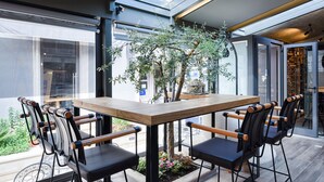 Un espace de restauration extérieur moderne, avec une table en bois et des chaises noires, entouré de murs en verre et offrant une vue sur la rue.
