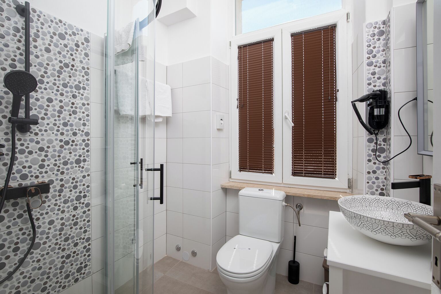 A bathroom with a glass shower enclosure, a toilet, a sink with a patterned basin, and a window with blinds.